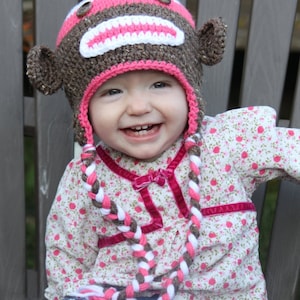 May include: A young child wearing a brown and pink monkey hat with a white pom-pom on top. The hat has a long braided strap that hangs down the front of the child's white floral dress. The child is smiling and sitting on a wooden chair.