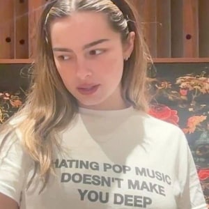 Puede incluir: Camiseta blanca con el texto negro "HATING POP MUSIC DOESN'T MAKE YOU DEEP". La persona que lleva la camiseta tiene el pelo largo y rubio ondulado y una diadema negra. El fondo tiene un diseño floral.