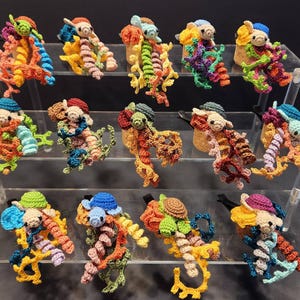 May include: A collection of colorful, handmade crochet sea creatures displayed on a tiered acrylic stand. Each creature features a unique combination of bright colors, including orange, blue, green, and yellow, with intricate details.