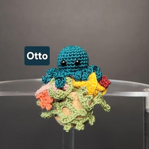 May include: A teal crocheted octopus with black eyes sits atop a colorful crocheted coral reef. The reef features yellow starfish, red and orange accents, and green and beige coral. The word "Otto" is displayed in a white font on a blue background.