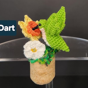 May include: A crochet hummingbird figurine with bright green wings and a light-colored chest, perched on a cork base. The bird is positioned near a yellow and orange flower and a white flower with a yellow center. The text Dart is visible.