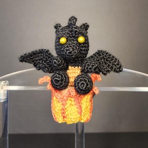 May include: A handmade, crocheted creature with a black body, wings, and a small head with yellow eyes. It is perched atop a yellow and orange crocheted pumpkin. The wings are spread, and the creature has a whimsical appearance.