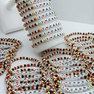 May include: A collection of beaded bracelets with the phrases "I LOVE PASTA" and "I LOVE CARBS" spelled out in white letter beads. The bracelets feature red, black, and gold beads, creating a colorful and playful design. The bracelets are displayed on a white stand.
