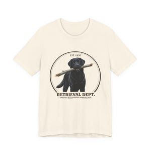May include: Cream-colored t-shirt featuring a black Labrador Retriever holding a stick in its mouth. The dog is inside a circular graphic with the text "RETRIEVAL DEPT. - OBJECT RECOVERY SPECIALIST" and "EST. 1830".