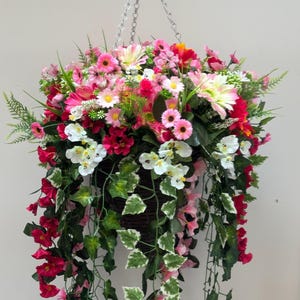 May include: A hanging basket filled with an assortment of artificial flowers in shades of pink, red, white, and yellow. Trailing green vines and leaves cascade down. The basket is suspended by a silver chain.