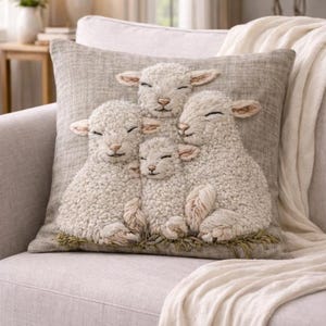 Puede incluir: Cojín cuadrado decorativo con un diseño bordado de cuatro ovejas blancas con detalles marrones claros, sobre un fondo gris neutro. El cojín está colocado en un sofá gris claro, con una manta blanca sobre el brazo.