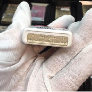 May include: A white Zippo lighter with the word "Marlboro" visible on the top. The bottom of the lighter is tan and has the Zippo logo, the text "BRADFORD, PA. MADE IN U.S.A.", and the number "18".