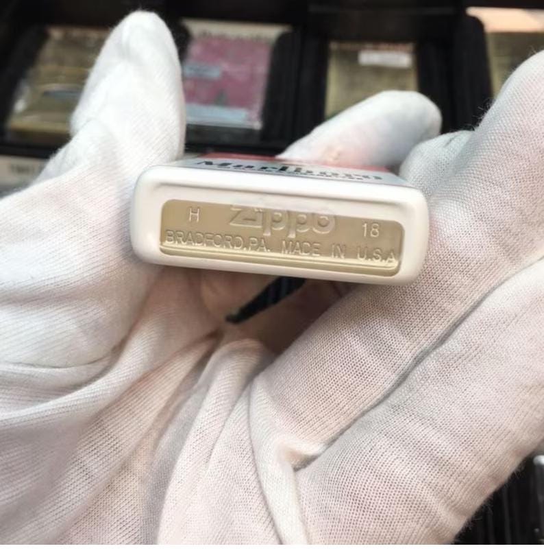 May include: A white Zippo lighter with the word "Marlboro" visible on the top. The bottom of the lighter is tan and has the Zippo logo, the text "BRADFORD, PA. MADE IN U.S.A.", and the number "18".