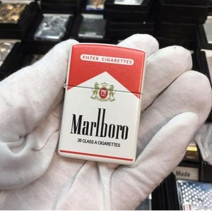 May include: A rectangular, white and red lighter with the Marlboro logo and text. The lighter is held in a gloved hand. The top of the lighter reads "FILTER CIGARETTES". The lighter is a classic design.