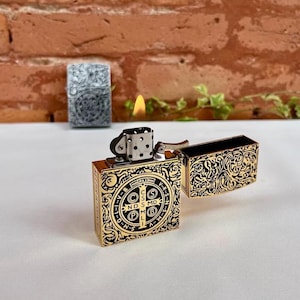 May include: Gold-colored metal lighter with intricate black filigree and a religious emblem. The lighter is open, revealing the interior. A second, silver-colored lighter is in the background. A flame is lit.