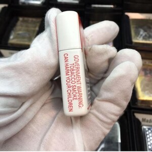 May include: A white and red rectangular object with the text "GOVERNMENT WARNING: TOBACCO SMOKE CAN HARM YOUR CHILDREN." The object is held in a gloved hand. The top of the object is white.