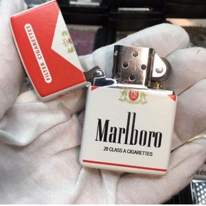 May include: A white and red Marlboro lighter with the brand name in black lettering. The lighter is open, revealing the interior. The top of the lighter has a silver hinge and a small wheel. The cigarette box is red and white.