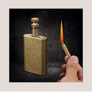 May include: A brass-colored, rectangular lighter with a screw-top mechanism. A lit match is held in the hand, showing a flame. The lighter has a textured surface and a vintage aesthetic.