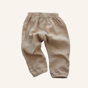Pode incluir: Uma calça bege clara, larga, feita de um tecido macio e texturizado. A calça tem cintura elástica e um caimento solto.