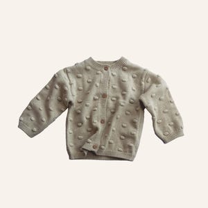 Puede incluir: Un cárdigan de punto beige con cierre de botones y un patrón texturizado. El cárdigan tiene mangas largas y un cuello redondo.