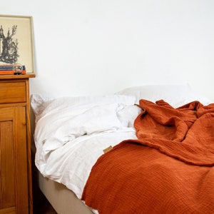 Puede incluir: Una cama blanca con una manta de color óxido. La cama está hecha con sábanas blancas y tiene una cabecera de madera. Un tocador de madera con un cajón y una puerta está en el fondo.