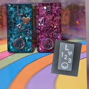 Pode incluir: Três peças retangulares de resina com glitter e enfeites. Uma é azul-turquesa com a letra "L", os números 1, 2 e 3 e um coração. Outra é rosa com a letra "R", os números 4, 5 e 6 e uma estrela. Uma peça cinza menor também está presente.