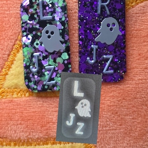 May include: Two rectangular resin charms featuring glitter and ghost motifs. The left charm has green and black glitter with the letters L, J, and Z. The right charm has purple glitter with the letters R, J, and Z. A small gray overlay shows the letters L, J, Z and a ghost.
