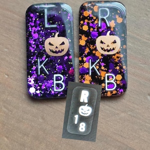 May include: Two rectangular resin charms featuring purple and orange glitter, each with a jack-o'-lantern graphic and the letters L, K, B or R, K, B. A smaller inset shows a rectangular charm with the letter R, a jack-o'-lantern, and the numbers 1 and 8.