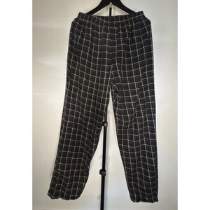 May include: Black and white checked trousers hanging on a black hanger. The trousers have a wide leg and an elasticated waistband. The checked pattern is made of thin white lines on a black background. The trousers are made of a lightweight fabric.