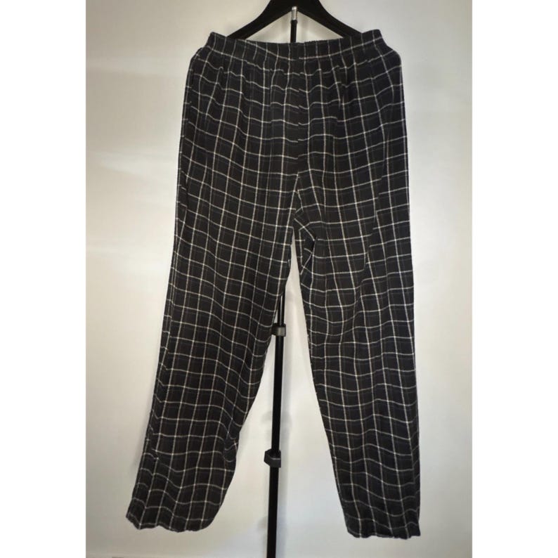 May include: Black and white checked trousers hanging on a black hanger. The trousers have a wide leg and an elasticated waistband. The checked pattern is made of thin white lines on a black background. The trousers are made of a lightweight fabric.