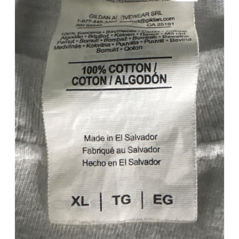 May include: Close-up of a white clothing tag with black text. The tag reads "100% COTTON / COTON / ALGOD&Oacute;N" and "Made in El Salvador". The tag also includes size indicators: XL, TG, and EG. The tag is attached to a gray fabric.