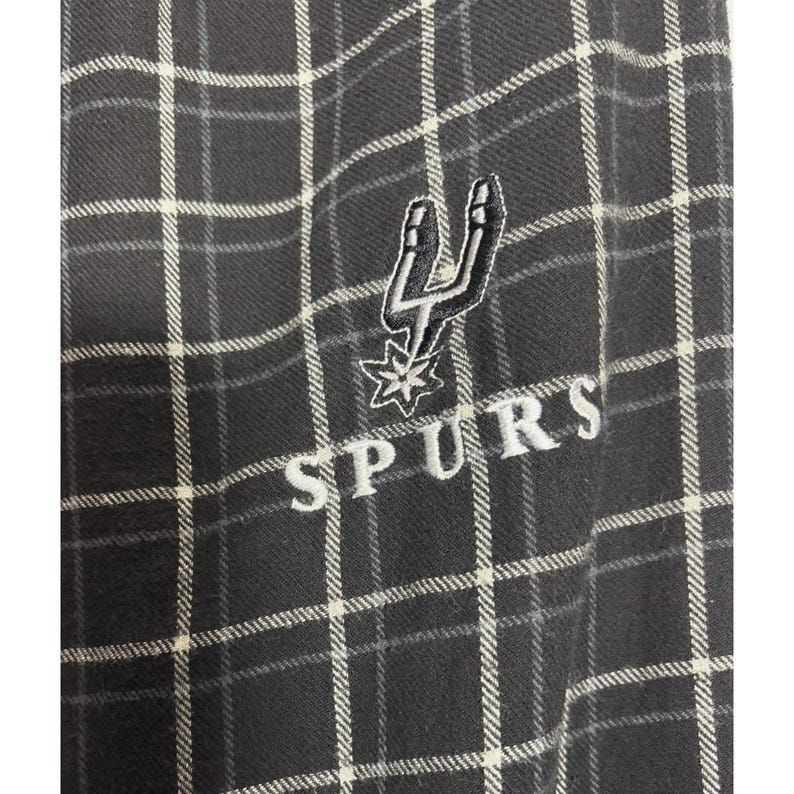 May include: A close-up of a black and white checked fabric with the word "SPURS" embroidered in white. The Spurs logo, a stylized spur and star, is also embroidered in white.