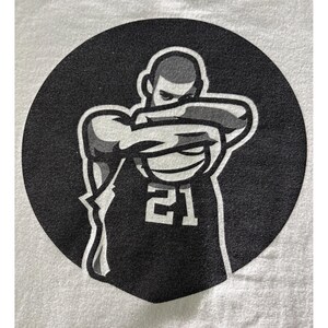 May include: A black and white graphic of a person in a jersey with the number 21, arms crossed in front of the face, set against a black circle. The image is printed on a white t-shirt.