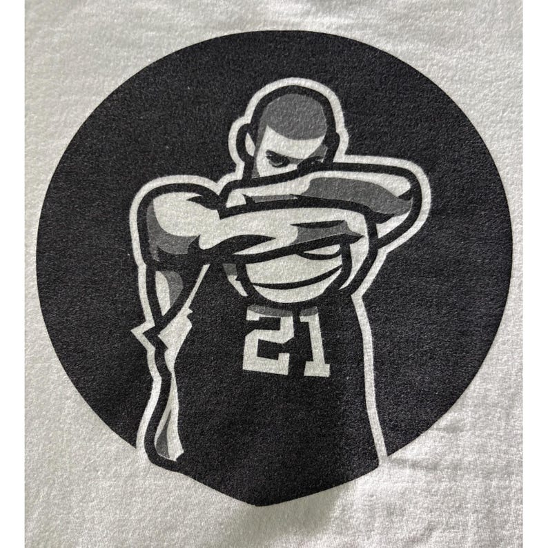 May include: A black and white graphic of a person in a jersey with the number 21, arms crossed in front of the face, set against a black circle. The image is printed on a white t-shirt.