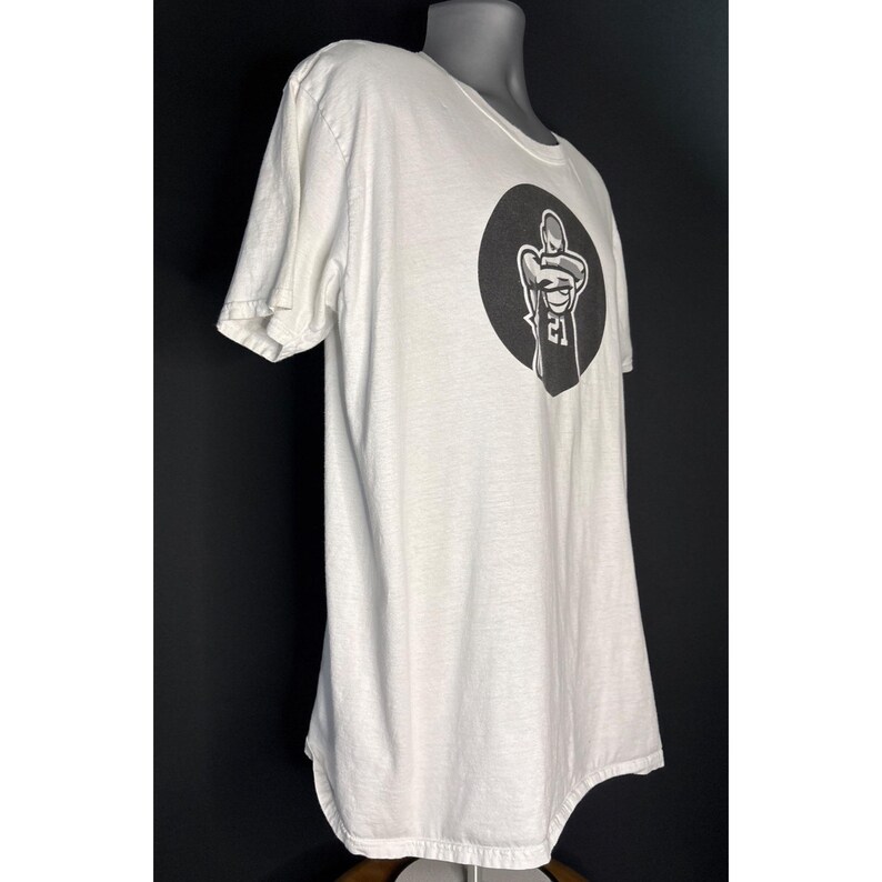 May include: White crew neck t-shirt with a black graphic on the front. The graphic is a circular design with a stylized figure holding a basketball. The shirt has short sleeves and a curved hemline.