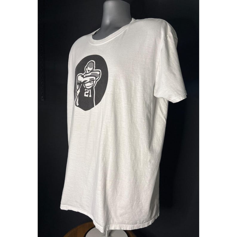 May include: White t-shirt with a black graphic of a basketball player in a circle, with the number 21 visible. The shirt has a crew neck and short sleeves. The shirt is displayed on a mannequin.
