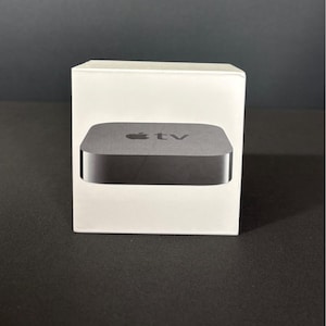Può includere: Una scatola bianca con un dispositivo Apple TV visibile. L'Apple TV è di colore grigio scuro con il logo Apple e la parola "tv" stampata sul dispositivo. La scatola è appoggiata su una superficie scura.