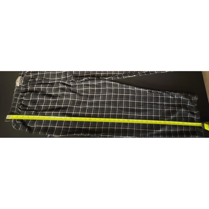 May include: Black and white plaid pajama trousers with a yellow measuring tape laid across them. The trousers have a button closure and a drawstring waist. The trousers measure approximately 101 cm in length.