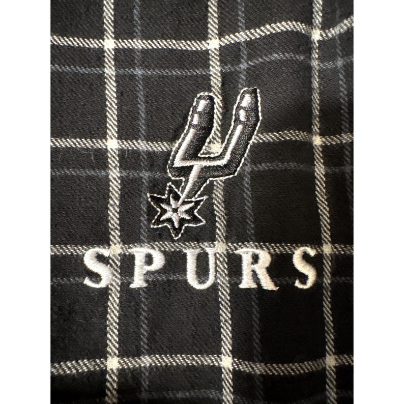 May include: Black and white checked fabric with an embroidered San Antonio Spurs logo and the word "SPURS" in white. The logo features a silver spur and a star.