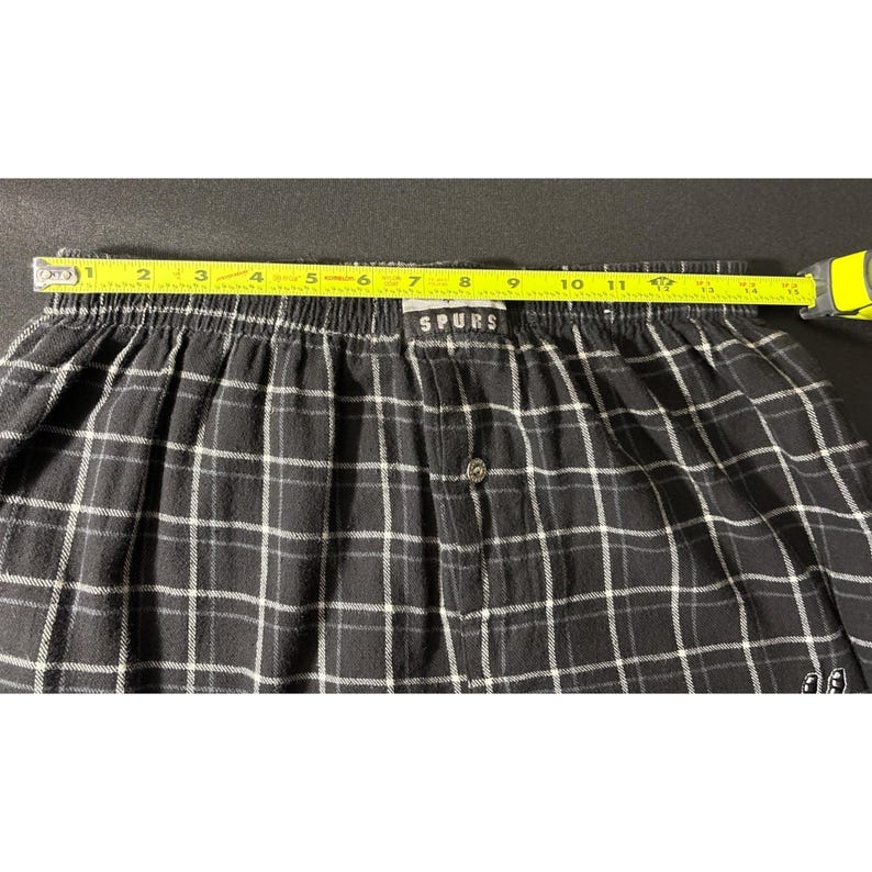 May include: Black and white checked boxer shorts with the word "SPURS" on the waistband. The shorts are measured with a yellow tape measure, showing a length of approximately 30 cm.