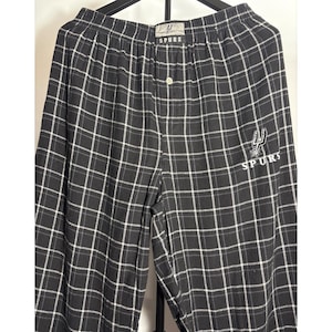 May include: Black and white checked pajama trousers with the word "SPURS" printed on the waistband and leg. The trousers have a button closure and a logo of a spur and the word "SPURS" on the leg. The trousers are made of a soft, comfortable material.