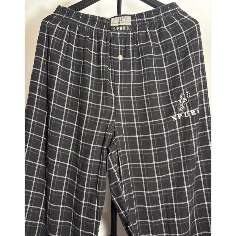 May include: Black and white checked pajama trousers with the word "SPURS" printed on the waistband and leg. The trousers have a button closure and a logo of a spur and the word "SPURS" on the leg. The trousers are made of a soft, comfortable material.