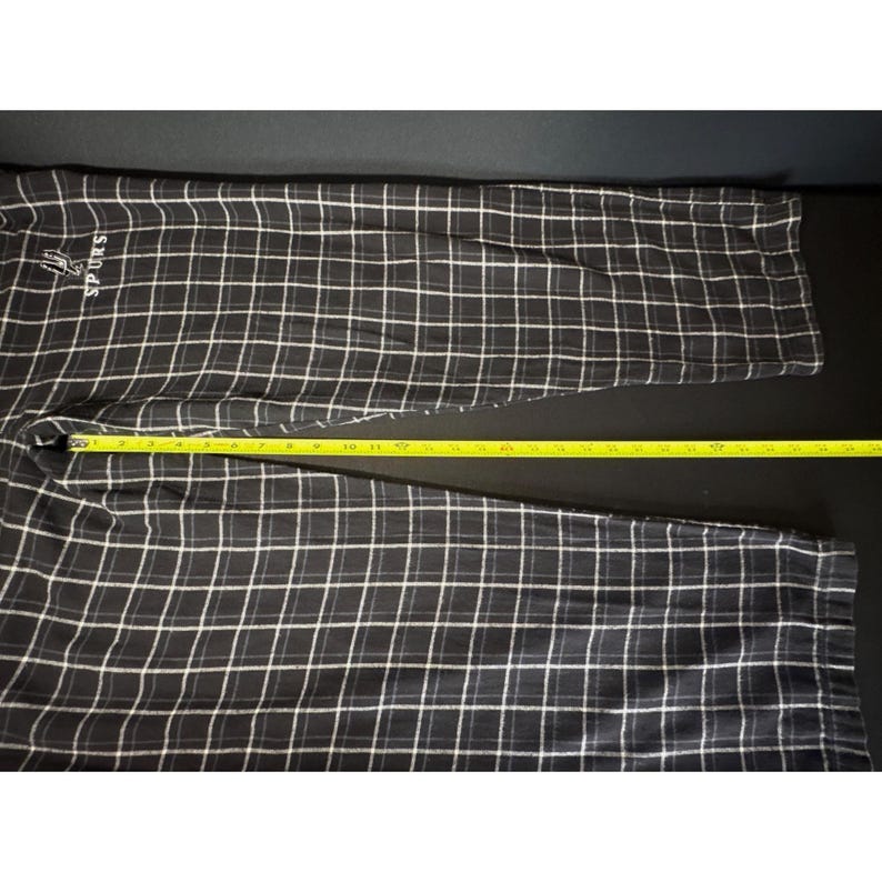 May include: Black and white checked pajama trousers with the word "SPURS" embroidered in white. The trousers are measured with a yellow measuring tape. The trousers are made of a soft material.
