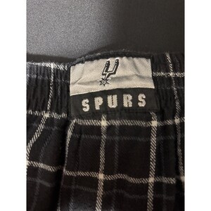 May include: Black and white checked pajama trousers with the word "SPURS" printed on a tag. The tag also features a team logo. The trousers are made of a soft, comfortable material.