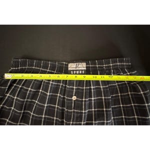 May include: Black and white plaid boxer shorts with a button closure. The waistband has the word "SPURS" printed on it. The shorts measure approximately 30 cm across.