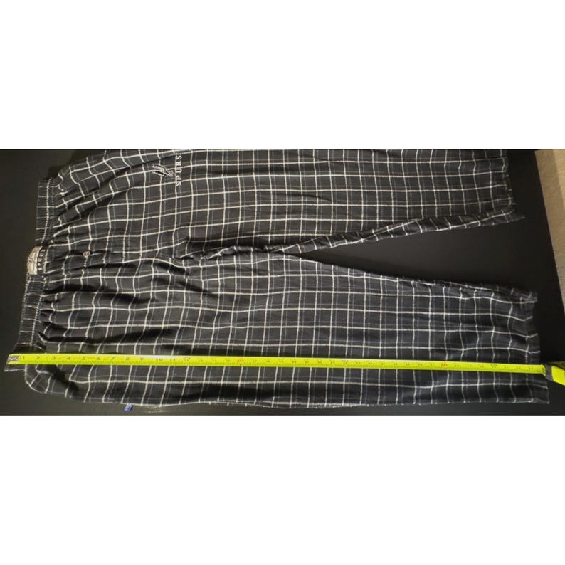 May include: Black and white checked pajama trousers with an elasticated waistband. The trousers have a white grid pattern over a black background. A yellow measuring tape is laid across the trousers. The trousers have the word "SPORTS" printed on them.