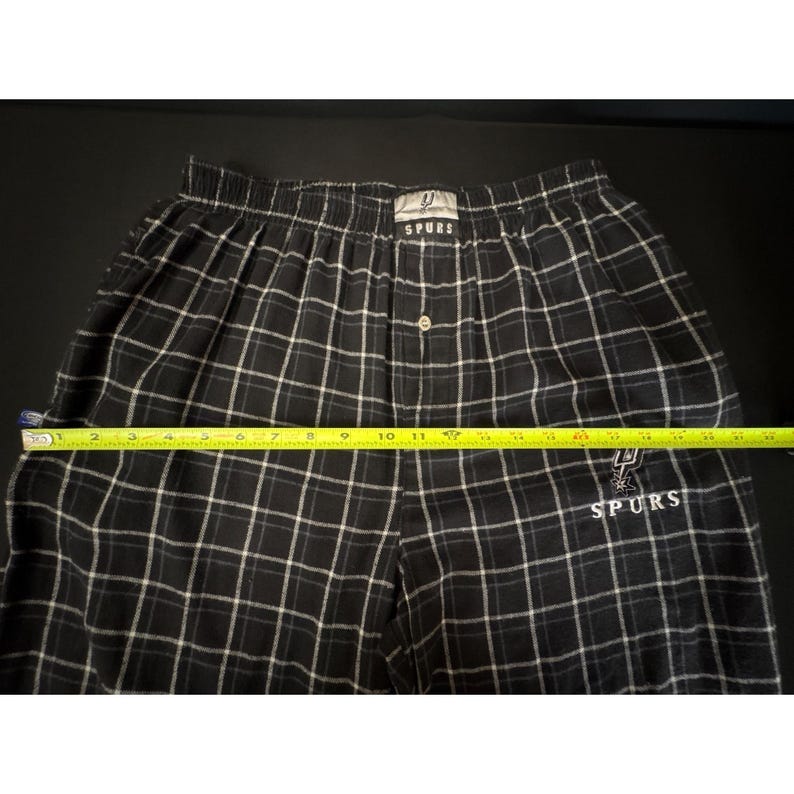 May include: Black and white plaid pajama trousers with the San Antonio Spurs logo embroidered on the leg. The trousers have a button closure and an elastic waistband. The trousers measure approximately 30 cm across the waist.