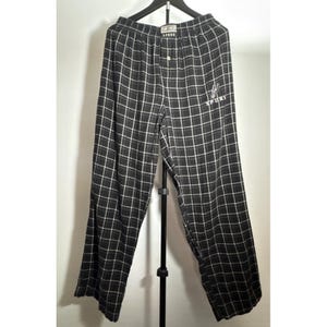 May include: Black and white checked pajama trousers hanging on a black hanger. The trousers feature a button closure and an embroidered design with the word "SPORT". The check pattern is a grid of black lines on a white background.