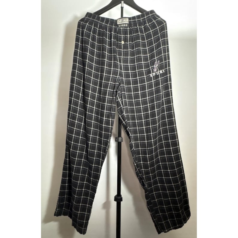 May include: Black and white checked pajama trousers hanging on a black hanger. The trousers feature a button closure and an embroidered design with the word "SPORT". The check pattern is a grid of black lines on a white background.