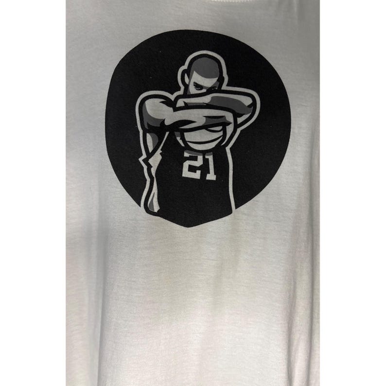May include: White t-shirt featuring a black and white graphic of a basketball player in a jersey with the number 21. The player is depicted with arms crossed over a basketball, set against a black circular background.