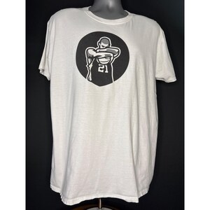 May include: White t-shirt with a black and white graphic of a basketball player inside a black circle. The player is wearing a jersey with the number 21. The shirt has short sleeves and a crew neck.