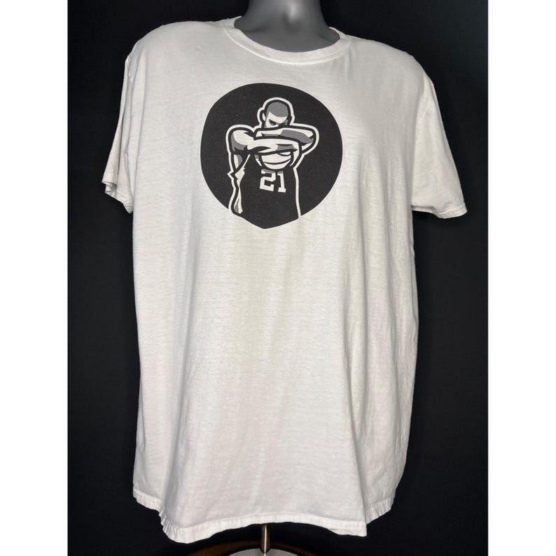 May include: White t-shirt with a black and white graphic of a basketball player inside a black circle. The player is wearing a jersey with the number 21. The shirt has short sleeves and a crew neck.