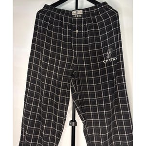 May include: Black and white checked pajama trousers with the word "SPURS" embroidered on the right leg. The trousers have a button closure and a small logo near the waistband.