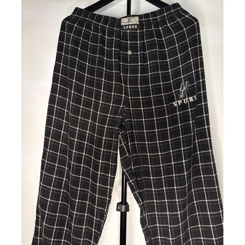 May include: Black and white checked pajama trousers with the word "SPURS" embroidered on the right leg. The trousers have a button closure and a small logo near the waistband.
