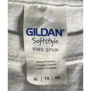 May include: Close-up of a white Gildan Softstyle t-shirt tag. The tag includes the brand name, fabric type, and country of origin. The tag also shows the size XL, TG, and EG.
